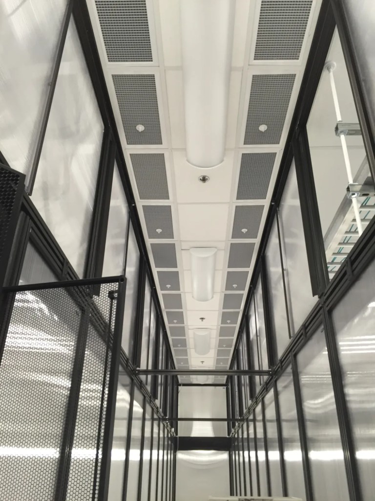Interior view of a long corridor featuring a grid ceiling with integrated lights and ventilation grilles, surrounded by black-framed transparent walls.