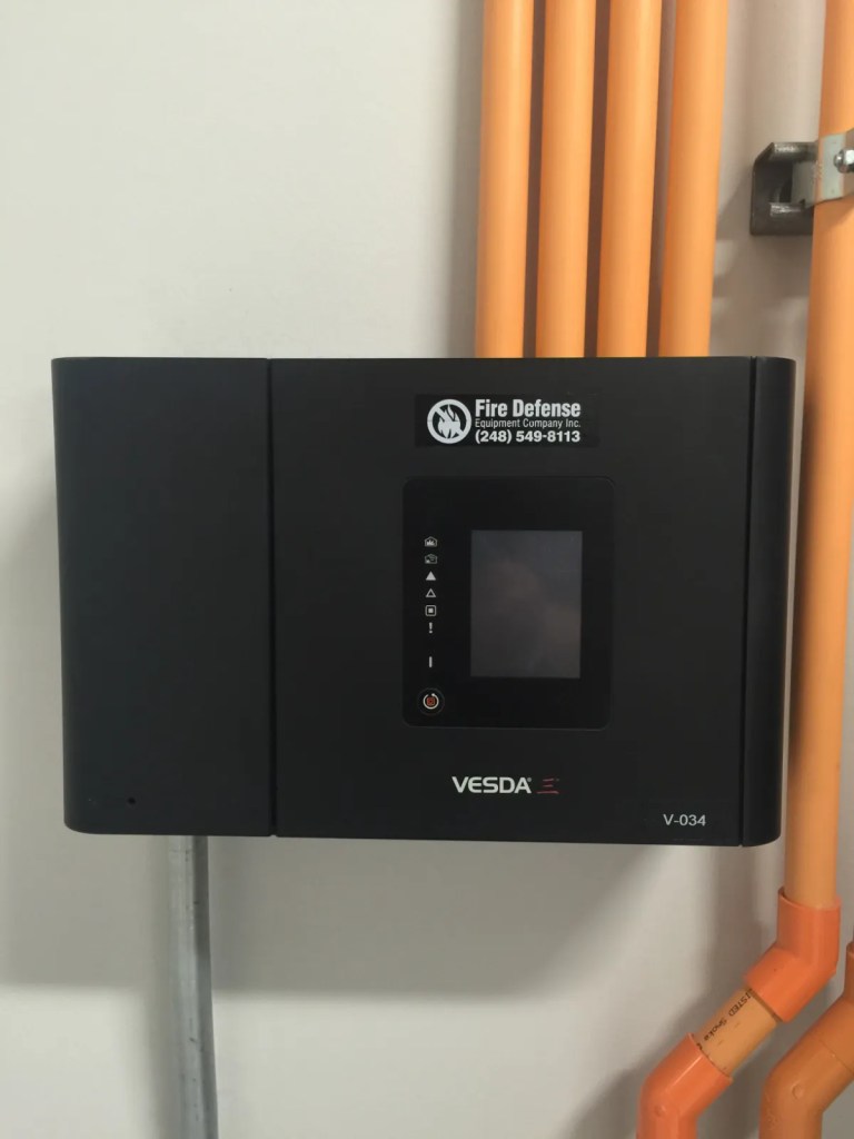 Fire alarm control panel with digital display mounted on a wall, accompanied by orange piping.