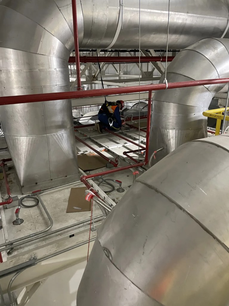 Worker in safety gear inspecting large metallic ducts and piping in an industrial setting.