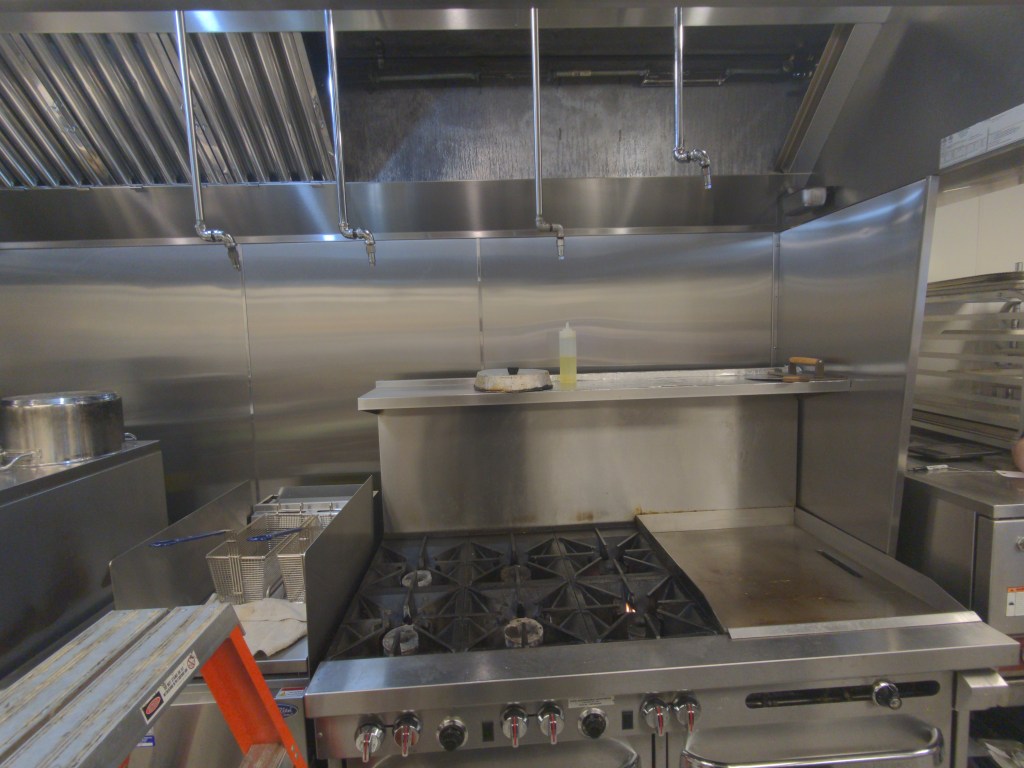A modern commercial kitchen featuring stainless steel surfaces, a gas range with burners, and cooking equipment including a grill and deep fryer. A bottle of oil is placed on the countertop.