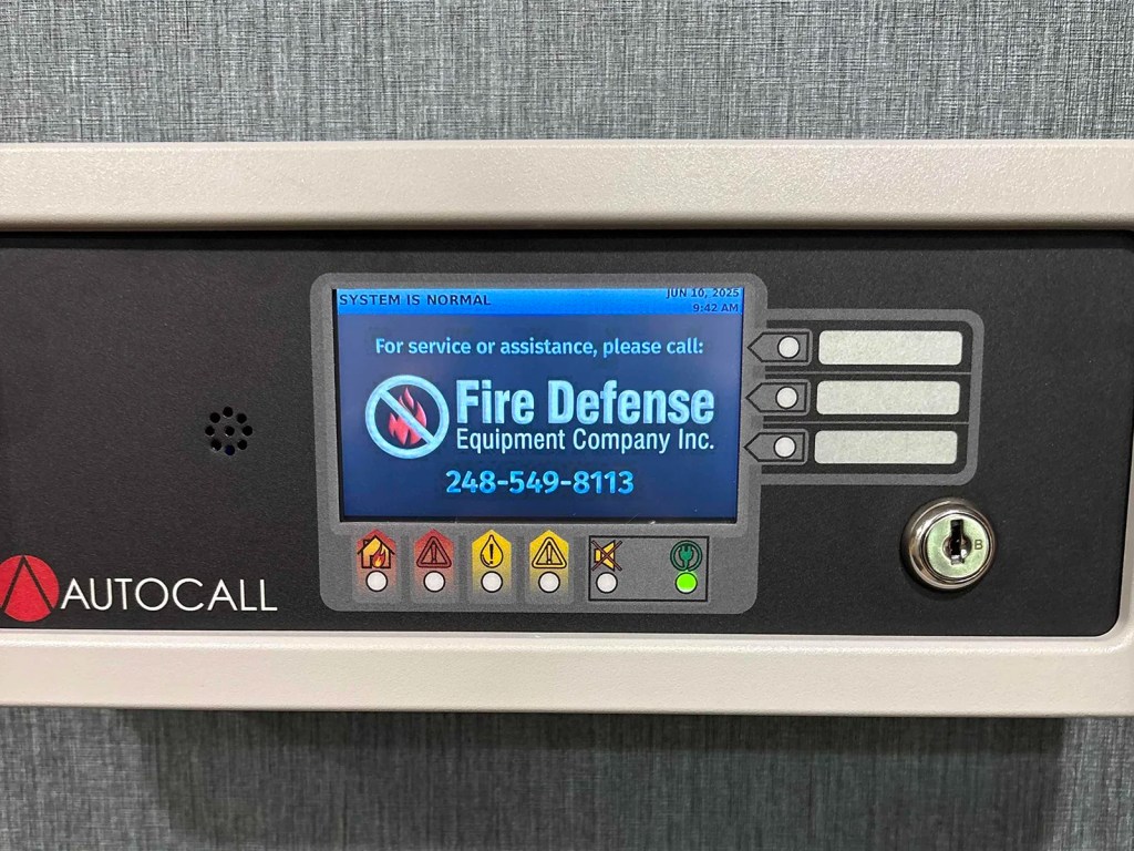 Control panel display labeled 'AUTOCALL' indicating 'SYSTEM IS NORMAL' with a contact for Fire Defense Equipment Company and a phone number.