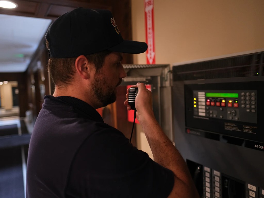 A man wearing a cap is speaking into a radio while standing next to a fire alarm control panel.