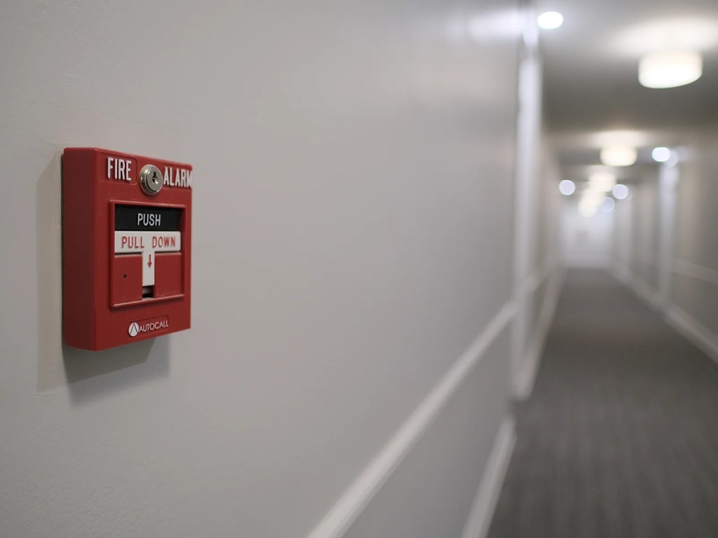 A close-up of a red fire alarm switch mounted on a wall in a hallway, with an out-of-focus background showing the corridor.