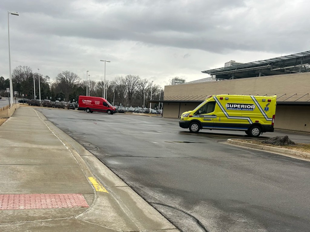 An ambulance with yellow and blue markings parked in a mostly empty lot beside a red delivery van, under a cloudy sky.